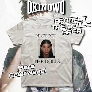 Może przedstawiać: Jasnoszara koszulka z napisem "PROTECT THE DOLLS" i zdjęciem osoby. Koszulka ma również napis "More Colorways!" i "ARCA". Słowo "OKINOWO" znajduje się na górze.