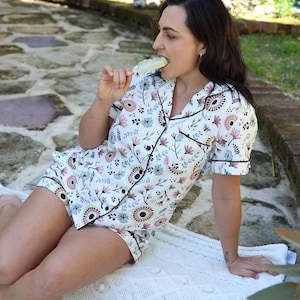 May include: White pajama set with a floral pattern in pink, blue, and brown. The short-sleeved top and shorts have black trim. The person is sitting on a white blanket.