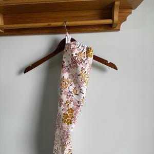 May include: A long, cylindrical fabric bag featuring a floral pattern with mustard yellow, dusty pink, and dark brown flowers on a white background. The bag hangs from a wooden hanger and includes a small white tag near the top opening.
