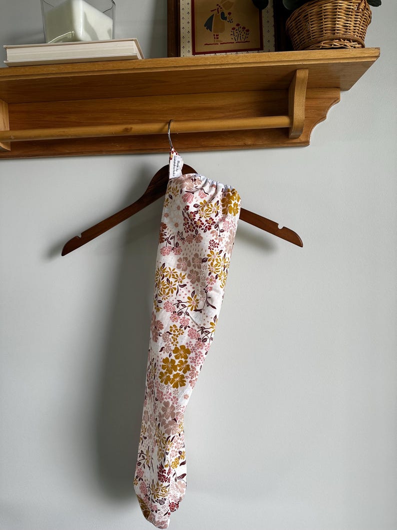May include: A long, cylindrical fabric bag featuring a floral pattern with mustard yellow, dusty pink, and dark brown flowers on a white background. The bag hangs from a wooden hanger and includes a small white tag near the top opening.