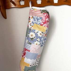 May include: A cylindrical fabric bag featuring a patchwork design of various floral, striped, and gingham patterns in shades of red, blue, yellow, and white. The bag has a white fabric top edge and a white hanging loop attached to the upper rim.