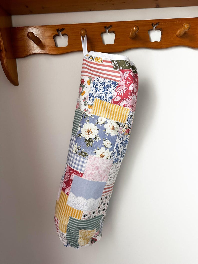 May include: A cylindrical fabric bag featuring a patchwork design of various floral, striped, and gingham patterns in shades of red, blue, yellow, and white. The bag has a white fabric top edge and a white hanging loop attached to the upper rim.