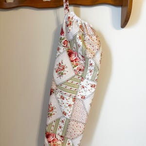 May include: A fabric bag with a patchwork design featuring various floral patterns in shades of red, green, and white. The bag has a gathered top opening and a fabric loop for hanging, constructed from multiple triangular and rectangular fabric panels.