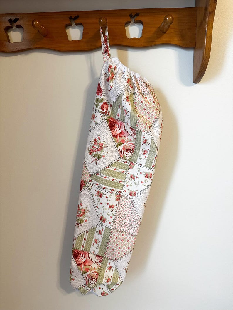 May include: A fabric bag with a patchwork design featuring various floral patterns in shades of red, green, and white. The bag has a gathered top opening and a fabric loop for hanging, constructed from multiple triangular and rectangular fabric panels.