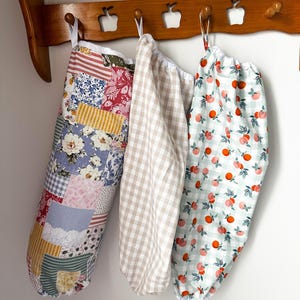 May include: Three fabric storage bags with elastic openings hanging from a wooden rack. The left bag features a multi-colored patchwork floral pattern. The center bag has a beige and white gingham check. The right bag displays an orange fruit print.