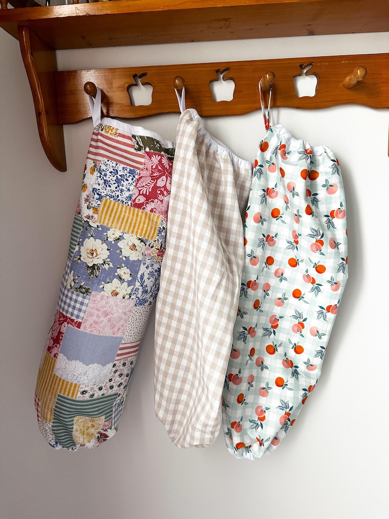 May include: Three fabric storage bags with elastic openings hanging from a wooden rack. The left bag features a multi-colored patchwork floral pattern. The center bag has a beige and white gingham check. The right bag displays an orange fruit print.