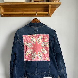 May include: A dark blue denim jacket with a quilted patch on the back. The patch features a pink star design surrounded by a floral pattern on a pink background. The jacket is hanging on a wooden hanger.