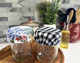 May include: Two clear glass Ball jars with wide mouths, each topped with a fabric lid. One lid features a blue floral pattern, the other a black and white checkered design. The jars sit on a round wooden cutting board.