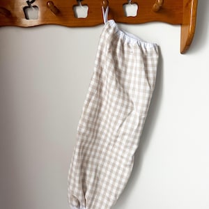 May include: A hanging fabric storage bag featuring a light beige and white gingham check pattern. The bag has a gathered white elastic opening at the top and a matching gathered white elastic opening at the bottom, designed for vertical storage.