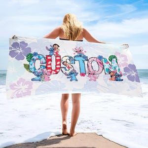 May include: A white beach towel with the word "CUSTOM" in colourful letters, featuring cartoon characters and floral designs. The towel is held up against a blue sky and ocean background.