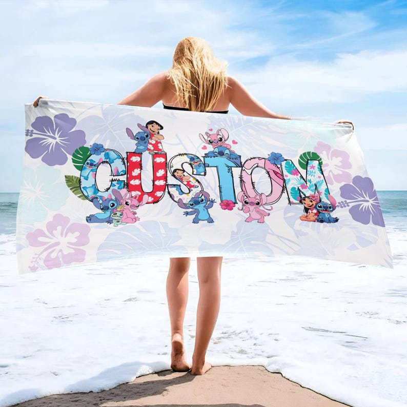 May include: A white beach towel with the word "CUSTOM" in colourful letters, featuring cartoon characters and floral designs. The towel is held up against a blue sky and ocean background.
