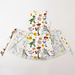 Op de afbeelding: Witte mouwloze jurk met een kleurrijk patroon van Toy Story-personages, waaronder Woody, Buzz Lightyear en Rex. De jurk heeft een uitlopende rok en bijpassende stoffen banden. Het ontwerp bevat sterren en het Toy Story-logo.