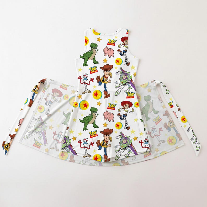 Op de afbeelding: Witte mouwloze jurk met een kleurrijk patroon van Toy Story-personages, waaronder Woody, Buzz Lightyear en Rex. De jurk heeft een uitlopende rok en bijpassende stoffen banden. Het ontwerp bevat sterren en het Toy Story-logo.