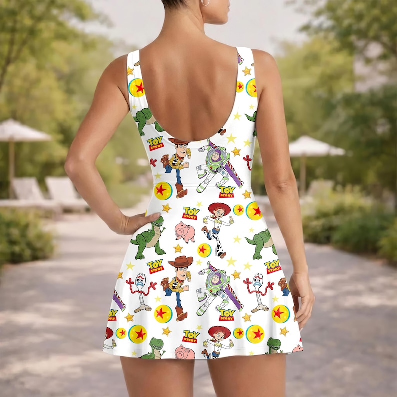 Op de afbeelding: Witte mouwloze badjurk met een herhalend patroon van Toy Story-personages, waaronder Woody, Buzz Lightyear en Rex. De jurk heeft een ronde rug en een uitlopende rok, met gele sterren en balafbeeldingen.