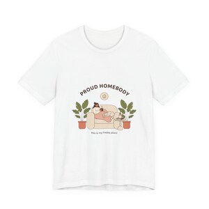May include: White t-shirt with a graphic design. The design features a person relaxing on a couch with a cat, surrounded by plants. The text "PROUD HOMEBODY" and "this is my happy place" are also included in the design.