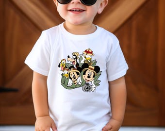 May include: A white t-shirt with a colourful graphic of Disney characters, including Mickey Mouse, Donald Duck, and Goofy. The characters are in a jungle setting. The child is wearing sunglasses and brown trousers.