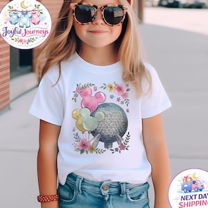 May include: A young person wearing a white t-shirt with a colorful graphic of a sphere, Mickey Mouse balloons, and flowers. The person is also wearing a pink cap and sunglasses. The shirt has the text "Joyful Journeys".
