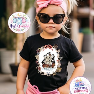 May include: A young child wearing a black t-shirt with a pirate ship design, pink pants, and a pink bow headband. The shirt features a detailed illustration of a pirate ship with mouse ears on the sails. The child is also wearing black sunglasses.