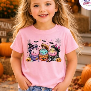 May include: A light pink short-sleeve t-shirt featuring a graphic print of three colorful, smiling dumpling characters in open containers, decorated with Halloween elements like a witch hat, bats, spiderwebs, and small pumpkins.