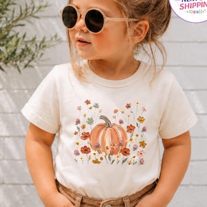 May include: A cream-colored short-sleeve t-shirt featuring a central graphic of an orange pumpkin surrounded by a variety of small, colorful wildflowers in shades of red, yellow, and green. The shirt has a crew neckline and a standard fit.