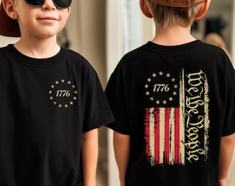 May include: Black t-shirt with the text "1776" and stars on the front, and a distressed American flag design with "We the People" on the back. The shirt is worn by a child.