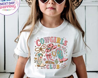 May include: A white short-sleeve t-shirt featuring a colourful graphic print with the text COWGIRLS CLUB, a lasso, a pink cowboy hat, a horseshoe, a pink bow, a belt, a cactus, and two cowboy boots decorated with floral patterns and stars.