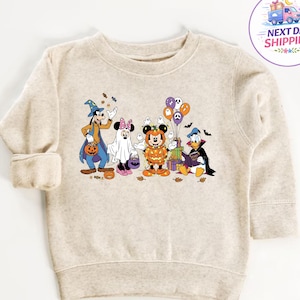 May include: A beige long-sleeve sweatshirt featuring a graphic print of Disney characters dressed in Halloween costumes, including Goofy as a wizard, Minnie as a ghost, Mickey as a pumpkin, and Donald as a vampire, surrounded by balloons.