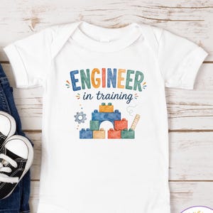 May include: A white short-sleeved baby bodysuit featuring the text Engineer in training in colorful letters above a graphic of stacked building blocks, a small gear, and a ruler. The garment has a round neckline and three metal snaps at the bottom.