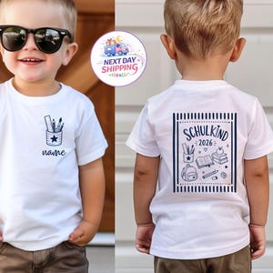 May include: A white short-sleeved t-shirt featuring a small dark blue graphic of a pencil holder on the front chest with the word name below it. The back displays a large dark blue graphic with the text SCHULKIND 2026, school supplies, and a backpack.