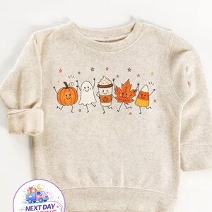 May include: A heathered beige long-sleeve sweatshirt featuring a central graphic of five cartoon characters: an orange pumpkin, a white ghost, a coffee cup, an orange maple leaf, and a candy corn, all with smiling faces and small stars.