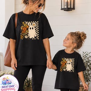 May include: Two black t-shirts featuring a graphic print of a white ghost surrounded by orange, yellow, and white flowers against a checkered background. The design includes a cluster of daisies and autumn-toned blossoms arranged beside the ghost.
