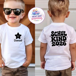 May include: A white short-sleeved t-shirt featuring a black star graphic with a smiling face and the name Daniel on the front chest. The back of the shirt displays the black text SCHUL KIND 2026 in a bold, wavy font with small star accents.