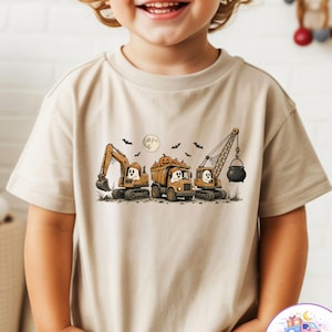 May include: A beige short-sleeve t-shirt featuring a graphic print of three construction vehicles, including an excavator, a dump truck filled with pumpkins, and a crane lifting a black cauldron, all operated by white ghosts under a full moon.