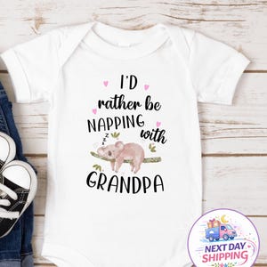 May include: White baby onesie with the text "I'd rather be napping with Grandpa" in black script. A sleeping koala illustration is on the onesie. The onesie is next to a pair of black and white baby shoes and blue jeans.