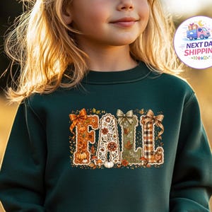May include: A dark green long-sleeve sweatshirt featuring the word FALL in large, decorative letters. Each letter is filled with autumn-themed patterns including pumpkins, leaves, mushrooms, and plaid, accented with small bows at the top.