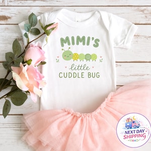 May include: A white short-sleeved baby bodysuit featuring a green caterpillar graphic and the text "MIMI'S little CUDDLE BUG" in green lettering, paired with a layered light pink tulle tutu skirt. The set is decorated with small pink hearts.