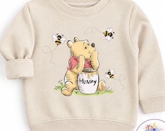 Könnte beinhalten: Ein beigefarbenes Langarm-Sweatshirt mit einer grafischen Illustration eines gelben Bären in einem roten Oberteil, der neben einem weißen Honigtopf mit der Aufschrift HUNNY sitzt, umgeben von drei kleinen fliegenden Bienen mit schwarz-gelben Streifen.