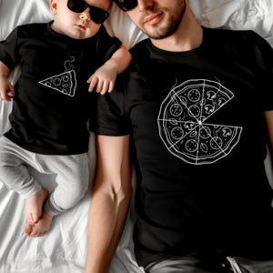 May include: Two black t-shirts with white pizza designs. One shows a whole pizza with a slice removed, the other a single slice. An adult and a baby, both wearing sunglasses, are wearing the shirts and lying on a white surface.