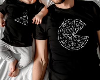 May include: Two black t-shirts with white pizza designs. One shows a whole pizza with a slice removed, the other a single slice. An adult and a baby, both wearing sunglasses, are wearing the shirts and lying on a white surface.