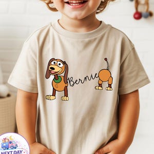 Op de afbeelding: Beige T-shirt met korte mouwen met een cartoonhondontwerp en de naam "Bernie". De hond is bruin, beige en wit met een groene kraag. De staart van de hond is ook zichtbaar.