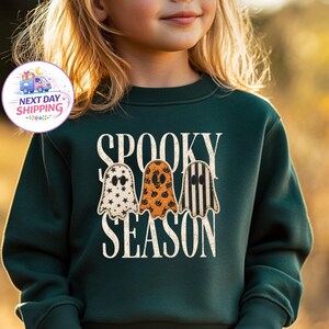 May include: A dark green long-sleeve sweatshirt featuring the text SPOOKY SEASON in white capital letters. Between the words are three ghost illustrations: one with black stars, one with orange pumpkins, and one with black and white vertical stripes.
