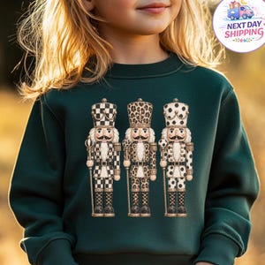 May include: A dark green long-sleeve sweatshirt featuring a graphic print of three nutcracker figures. The left nutcracker has a black and white checkered pattern, the center has a leopard print, and the right has a black and white polka dot pattern.