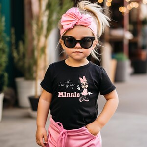 May include: A young child wearing a black t-shirt with the text "I'm this Minnie" and a Minnie Mouse graphic. The child is also wearing a pink headband, sunglasses, and pink pants. The image is taken outdoors.