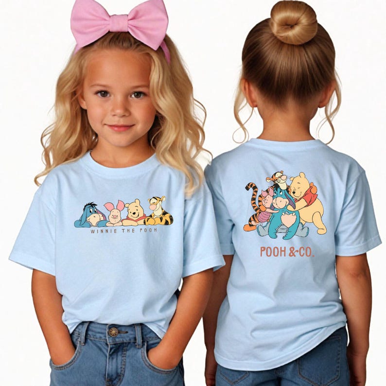 May include: Two young girls wearing light blue t-shirts featuring Winnie the Pooh characters. The front of the shirts display Eeyore, Piglet, Winnie the Pooh, and Tigger. The back of the shirts show the characters hugging with the text "POOH & CO."