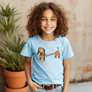 Op de afbeelding: Een lichtblauw T-shirt met een cartoonhond genaamd Bernie. De hond is bruin met een groene halsband en wordt in twee delen afgebeeld, met de naam Bernie ertussen geschreven. Het T-shirt wordt door een jong persoon gedragen.