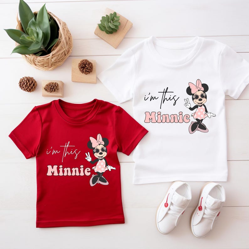 May include: Two short-sleeved t-shirts, one red and one white, featuring a Minnie Mouse graphic. The shirts have the text "I'm this Minnie" and are paired with white sneakers. Decorative plants and pinecones are in the background.