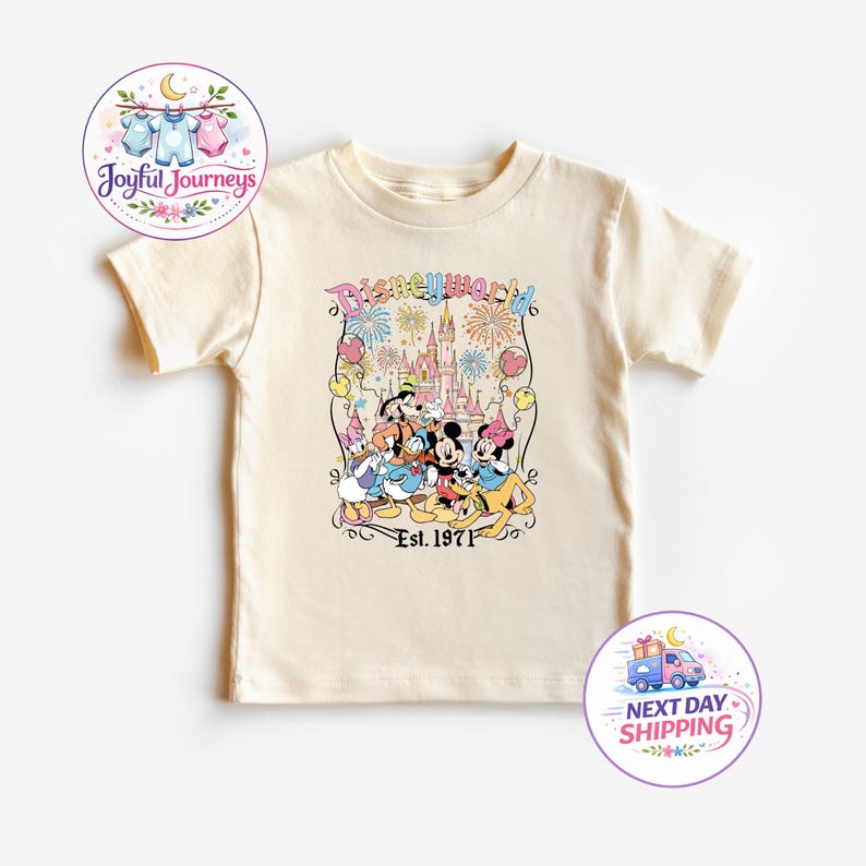 Op de afbeelding: Cr&egrave;mekleurig peuter-T-shirt met een kleurrijke afbeelding van Disney-figuren voor een kasteel, met de tekst "Disneyworld Est. 1971". Het shirt heeft korte mouwen en een ronde hals.