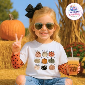 May include: A white short-sleeved t-shirt featuring a graphic print of nine assorted pumpkins with various patterns including leopard print, floral, and plaid. The shirt is layered over an orange long-sleeved shirt with black polka dots.