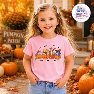 May include: A light pink short-sleeve t-shirt featuring a graphic print of four cartoon characters sitting inside orange jack-o'-lantern pumpkins, surrounded by small black bats. The characters include a pig, a bear, a tiger, and a donkey.
