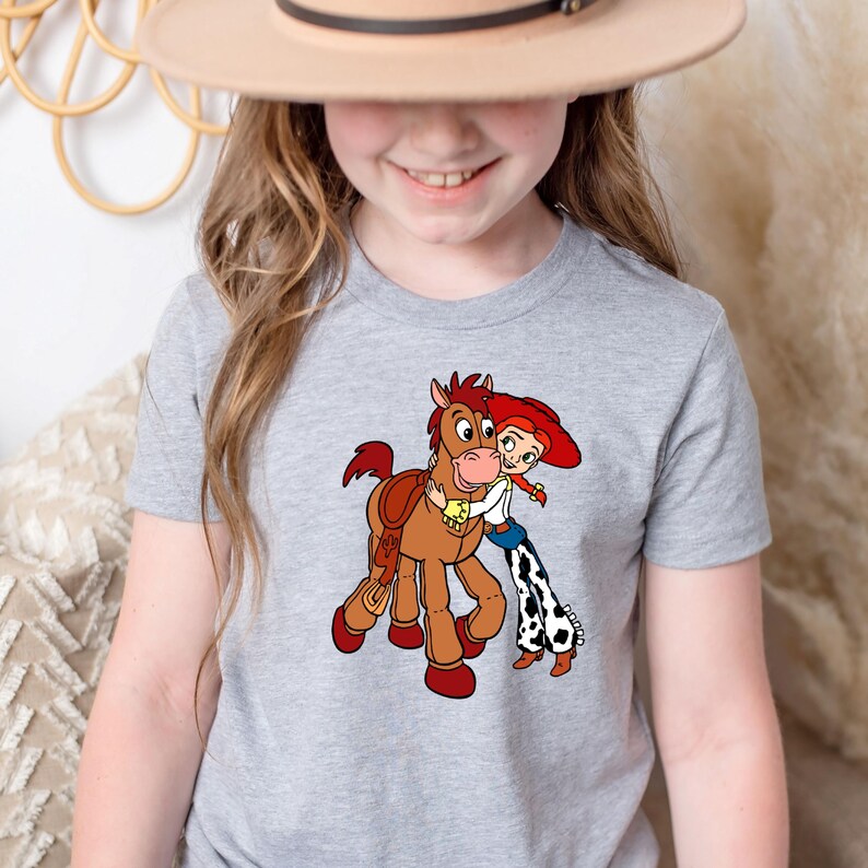 Puede incluir: Camiseta gris claro con un dibujo animado de un caballo marr&oacute;n abrazando a un personaje con sombrero rojo y vaqueros azules. El dibujo es de estilo caricatura. La camiseta es de material suave.